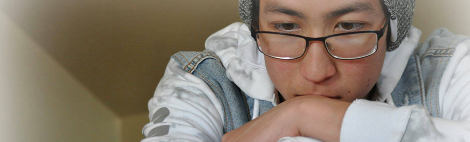 ID: photo of the head and shoulders of a teenage boy wearing eyeglasses, a hat and a white and jean sweatshirt, his arms are crossed and his chin/mouth are resting on his hands, he has a pensive look on his face