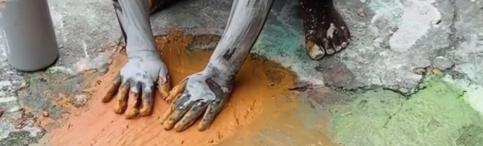 ID: photo of a child with chocolate skin playing in paints on an asphalt ground, her hands are covered in white and orange paint and there is orange, white, and green paint on the ground; the ground is cracked and textured; only the hands, forearms, and foot (with white pain splattered on it) of the child are visible in the photo