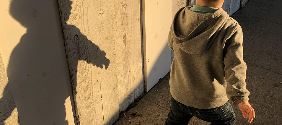 ID: photo of a boy running on a cement sidewalk wearing a grey sweatshirt and jeans, his blond hair is cut short and the photo only shows his back side with his shadow stark upon the fence beside him