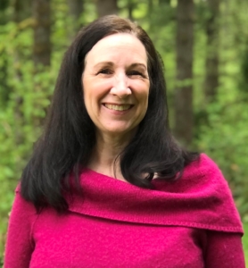 Image of author Kelly Rain Collin; white female with long straight dark hair, wearing a magenta sweater, smiling, standing in the woods