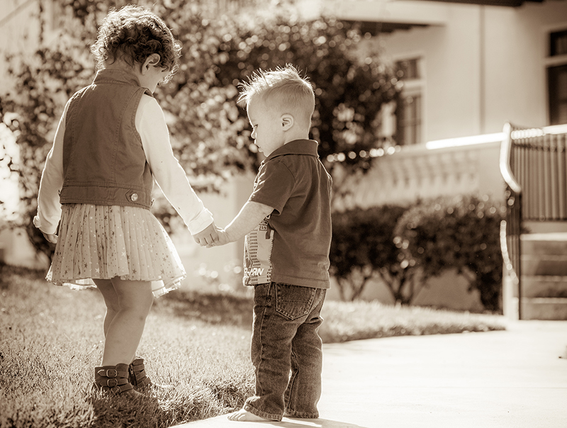 a sepia-tone image of a small blond 3-year old barefoot boy holds hands with a taller 3-year old dark-haired girl in a skirt outside on the edge of the sidewalk along the grass outside of a nicely-kept condominium complex on a bright sunny day.