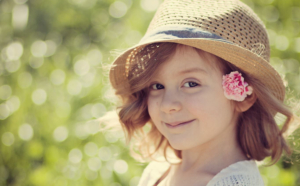 A young caucasian child with long red hair and a flower in her ear wearing a hat and a light sweater over a ruffled purple dress stands in a field of green grass. they are smiling sweetly, looking at the camera directly.