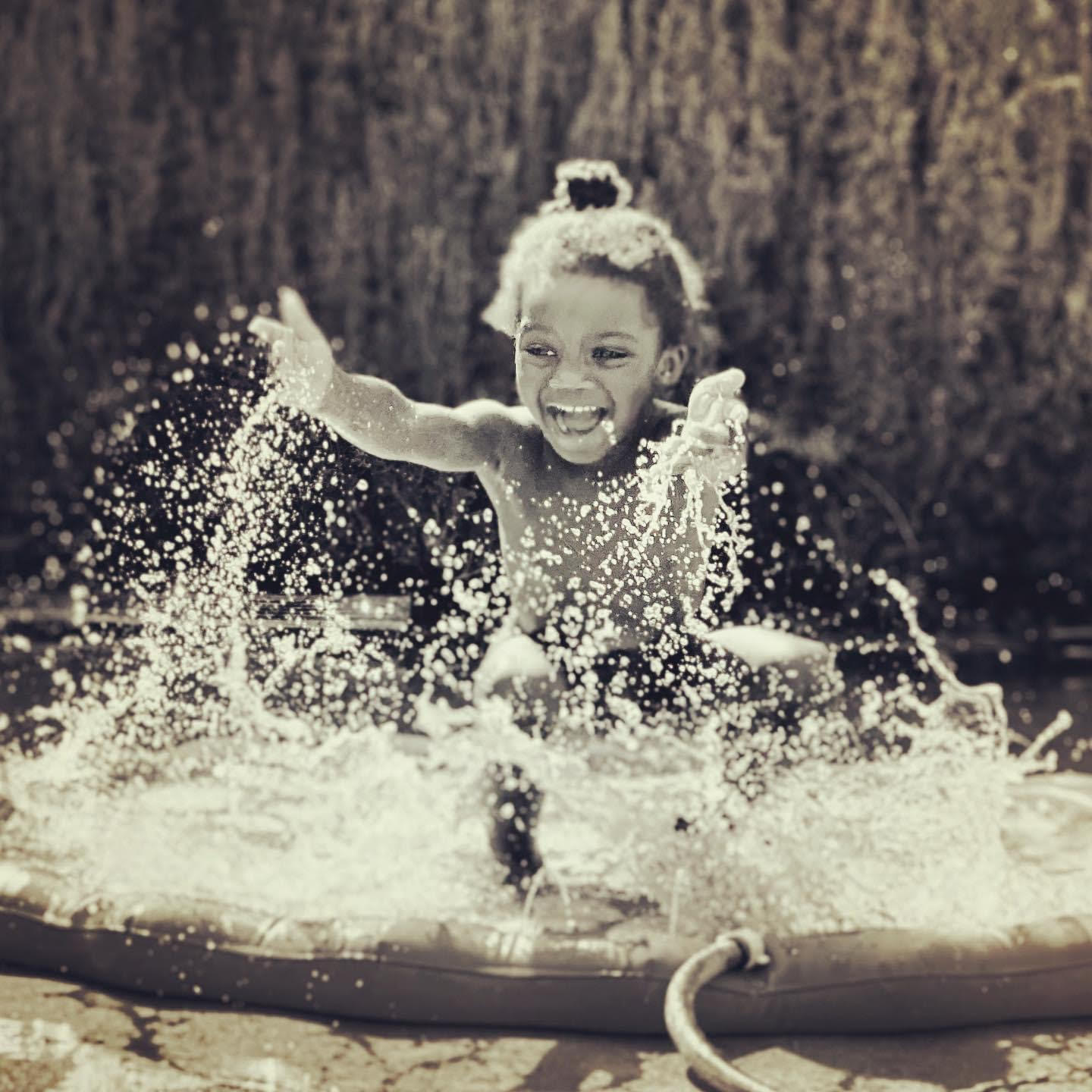 A young girl is sitting on a water-play mat filled with water and splashing it all around with a big open smile on her face. She is outside, with an indistinct background that is perhaps a fence or a hedge behind her.
