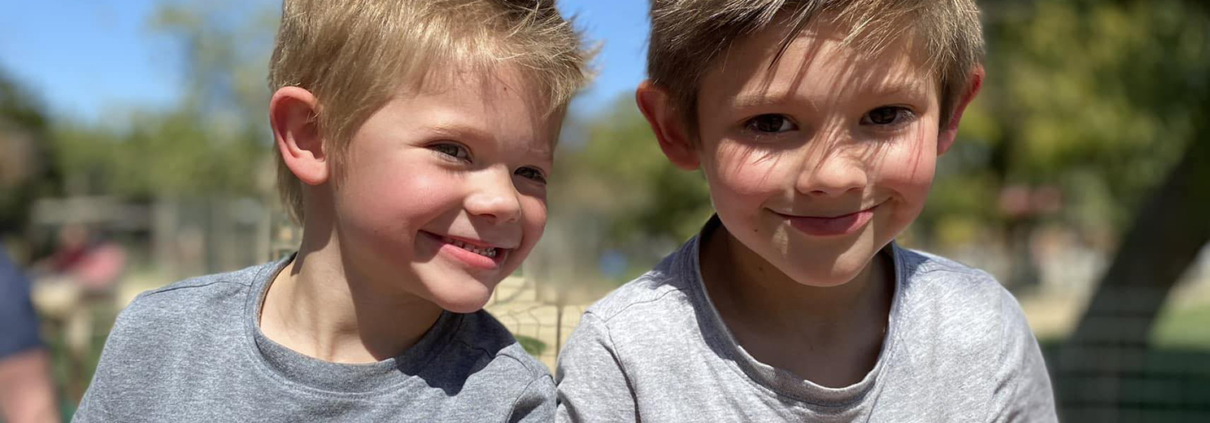 Two young brothers sit next to each other in a park. Both are wearing grey shirts and smiling. The shirt of one of the boys shows the ASL sign for friend.