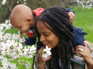 image of a mother, a woman of color with long black dreadlocks standing in the center-right of the foregrounds, leans to the left side of the image while holding a small child seated on the back of her neck. the child is leaning further to the left foreground, pulling some white flowers to their face to smell. the background is mostly a green grass field with some trees along the left margin going into the background.
