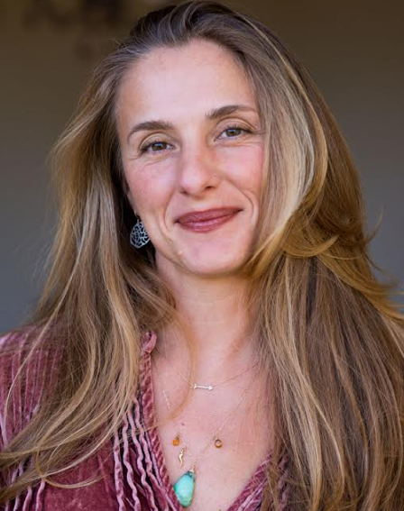image of Aleks Evenguelidi, LM, CPM. The image is a headshot, she is standing in front of neutral, out of focus background. She has long dark blonde hair. She is wearing medium silver patterned earrings, two thin necklaces with some stones, and a faded red top.