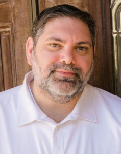 image of Dr. Elliot Berlin, a pregnancy-focused chiropractor based in Los Angeles. The image is a headshot, he is standing in front of an old wooden door. He has short brown hair, lightly graying at he temples, a salt-and-pepper beard, and is wearing a white button-down shirt.