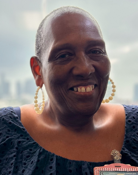 image of Toy R Hightower, CCBD, CLEC. The image is a headshot, she is standing in front of window, with the background of a cityscape in the distance and out of focus. She has very short cropped dark hair. She is wearing large, beaded loop earrings, and a dark blue top.