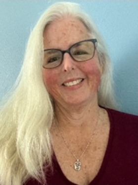 Image of author Rosemary Shulman; white female with long white hair worn straight, wearing a maroon top, she is smiling and the image shows her from the chest up, with a light blue background behind her