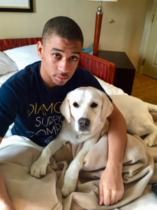 image of an African-American teen boy with short-cropped hair wearing a blue tshirt with indistinct writing on it and sitting on a bed wrapped in the comforter from the hips down. on his lap is a white/cream colored short-hair puppy with a dark nose and eyes. they are both looking into the camera at the viewer. they are on a fold-out couch-bed in a hotel room.