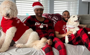 image of two teens with their service dogs, sitting on the sofa. the room appears to be a living room in a home, and the clothing and decorations suggest Christmas time. the teens (boy and a girl) are wearing red plaid christmas pajamas and the girl has a santa hat on. they are looking at the camera smiling. each has a white service dog on their lap.