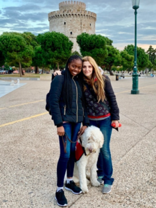 image of an African-American teen girl with her caucasian mother, outside in a public area with trees in teh background and in the distance a large, castle-looking tower. the ground they stand on is all a concrete walking area., with a small pool or pond in the midground to the left of the image. the people stand in the foreground dressed in warm clothes, holding each other as they pose, smiling to the camera. they have a white service dog in a red vest with them.