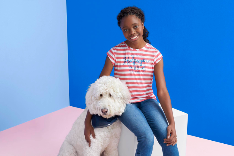 image of a young teen African-American girl sitting with her shaggy white service dog. she sits on a white cube with a flattened color background of pink, pale blue, and blue. she is wearing a red and white striped tshirt and bluejeans. the dog has a blue bandana tied around its neck.