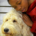 closeup image of a young African-American girl leaning into the picture from the right side so that we see her from the shoulders up ,wearing a soft red hoodie. she is snuggling into a large cream-colored golden doodle service dog with shaggy fur that we see from his neck up. the background is vague but appears to be part of a bedroom dresser.