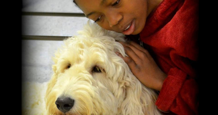 closeup image of a young African-American girl leaning into the picture from the right side so that we see her from the shoulders up ,wearing a soft red hoodie. she is snuggling into a large cream-colored golden doodle service dog with shaggy fur that we see from his neck up. the background is vague but appears to be part of a bedroom dresser.