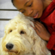 closeup image of a young African-American girl leaning into the picture from the right side so that we see her from the shoulders up ,wearing a soft red hoodie. she is snuggling into a large cream-colored golden doodle service dog with shaggy fur that we see from his neck up. the background is vague but appears to be part of a bedroom dresser.