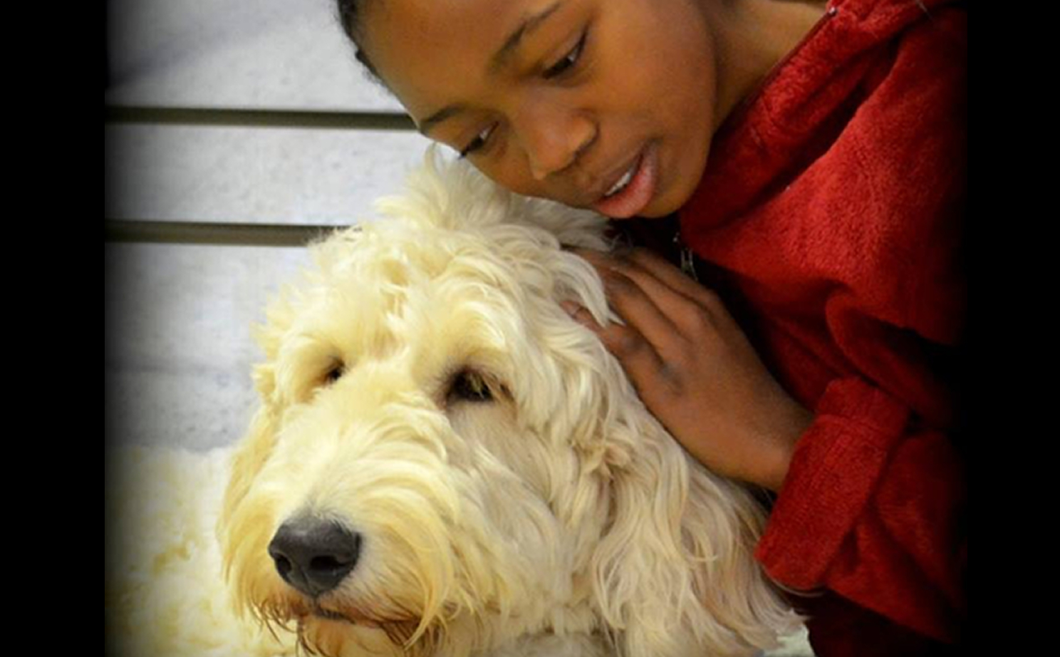 closeup image of a young African-American girl leaning into the picture from the right side so that we see her from the shoulders up ,wearing a soft red hoodie. she is snuggling into a large cream-colored golden doodle service dog with shaggy fur that we see from his neck up. the background is vague but appears to be part of a bedroom dresser.